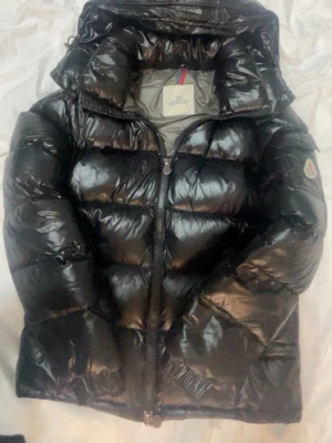 Moncler maya - Säljer en svart glansig dunjacka från Moncler med hög krage och huva. Jackan har en klassisk puffer-design, dragkedja framtill och Moncler-logga på ärmen. Perfekt för kalla vinterdagar och riktigt snygg streetstil.