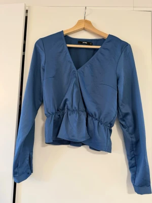 Blå omlottblus med volang - Snygg blå omlottblus med markerad midja och volang nedtill. Blusen har långa ärmar med knappdetalj vid ärmslut och ett mjukt, lätt glansigt tyg som ger en elegant känsla. Perfekt att styla med jeans eller kjol.