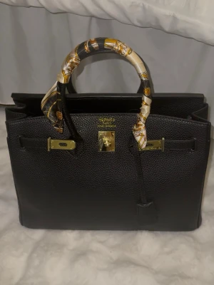 Svart handväska 35 cm - Lyxig svart Birkin-liknande handväska i präglat läder med guldfärgade metalldetaljer. Kommer med plasten innuti för att behålla formen. Silkes sjalen följer med som man ka ta bort om man önskar.