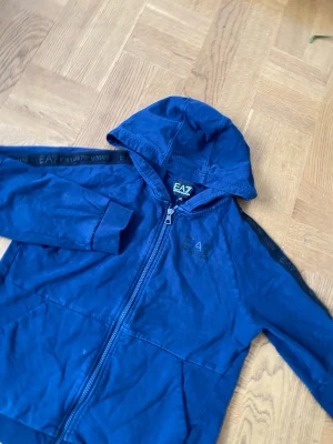 Blå hoodie från EA7 Emporio Armani - Snygg blå hoodie från EA7 Emporio Armani med dragkedja framtill och huva. Logga på bröstet och svarta detaljer längs ärmarna med EA7-tryck. Tillverkad i mjukt bomullsmaterial som känns skönt mot huden. Perfekt för en avslappnad och trendig look.