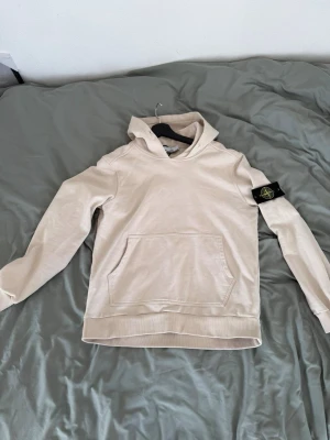 Beige hoodie från Stone Island - Snygg beige hoodie från Stone Island med klassisk patch på vänster ärm. Hoodien har en stor känguruficka framtill, ribbade muddar och en skön huva. Perfekt för en avslappnad och trendig stil. Tillverkad i mjuk bomull för extra komfort.