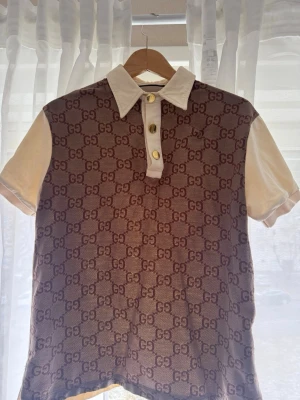 Beige GG-mönstrad henley från Gucci - Säljer en beige och brun henley från Gucci med ikoniskt GG-mönster över hela framsidan. Skjortan har korta ärmar, vit krage och knappslå med tre guldfärgade knappar. Perfekt för dig som vill sticka ut med lyxig stil.