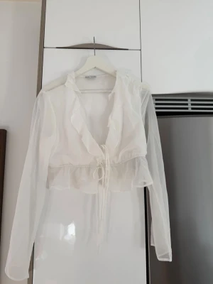 Vit omlott blus med volang och knyt - Säljer en vit, transparent omlott blus från NA-KD med volangdetaljer och knyt framtill. Blusen är croppad och har långa ärmar, perfekt för att skapa en trendig look. Materialet är lätt och luftigt, vilket gör den bekväm att bära.