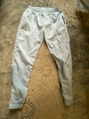 Ljusblå mjukisbyxor från Lacoste Sport - Säljer ett par ljusblå mjukisbyxor från Lacoste Sport med resår i midjan och vid bensluten. Byxorna har sidofickor och den klassiska Lacoste-loggan på benet. Perfekta för chill dagar eller träning. Materialet känns mjukt och bekvämt.