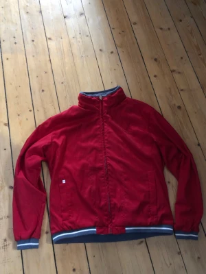 R H röd vindjacka från Umbro - Snygg röd vindjacka från Umbro med lodragkedja framtill och grå-vita detaljer vid muddar och nederkant. Jackan har hög krage, två sidofickor och Umb bologo på ryggen. Perfekt för dig som vill ha en sportig och stilren look.