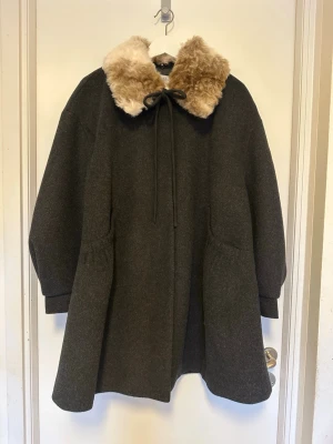 Mörkgrå kappa med fuskpäls - Säljer en oversized mörkgrå kappa med bred krage i beige fuskpäls och knytband framtill. Kappan har vida ärmar och en lös, A-linjeformad passform som ger en cool vintagekänsla. Perfekt för dig som vill ha en unik och mysig ytterrock till hösten eller vintern.
