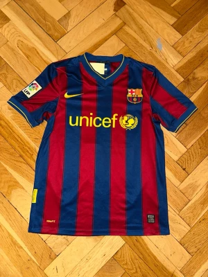 Retro Barcelon FC 2009-2010  - Säljer en klassisk FC Barcelona matchtröja från Nike med blå och röda vertikala ränder, gul Nike-logga och Unicef-tryck på bröstet. Tröjan har klubbmärke, La Liga-märke på ärmen och är tillverkad i lätt funktionsmaterial. Den är 2009-2010 säsong Tillhör legenden Puyol 