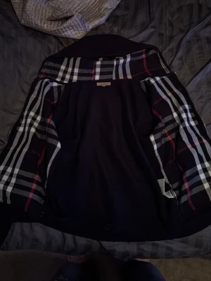 Svart ullkappa från Burberry - Stilren svart ullkappa från Burberry med klassiskt rutigt foder i svart, vitt och rött. Kappan har en rak passform och ikoniskt Burberry-mönster på insidan. Perfekt för dig som vill ha en tidlös och lyxig look allt är i perfekt skick snören t hoodien saknas