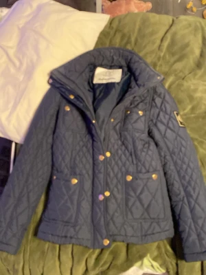 Marinblå quiltad jacka Chiara Forthi - Snygg marinblå quiltad jacka från Chiara Forthi Milano med guldfärgade knappar och fickor framtill. Jackan har hög krage, patch på ärmen och ett klassiskt rutmönster. Perfekt för dig som vill ha en stilren och trendig jacka till hösten.