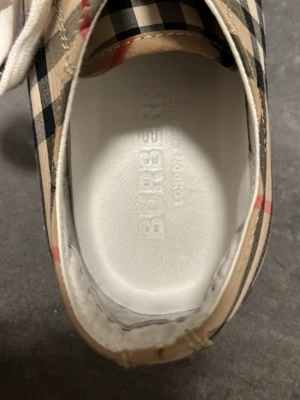 Burberry rutiga låga sneakers - Snygga låga sneakers från Burberry med det klassiska beige, svarta och röda rutmönstret. Skorna har vita snören och en vit innersula med Burberry-logga. Yttersulan är i gummi med hexagonmönster för bra grepp. Perfekta för dig som gillar lyxig streetstyle. Saknar tyvärr låda