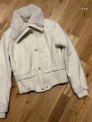 Guess Vit teddyjacka med stickad kant - Snygg vit teddyjacka med fluffig krage och stickad detalj längst ner. Jackan har tryckknappar framtill och två fickor. Perfekt för kalla dagar när du vill vara både varm och trendig.