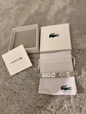 Silverfärgat armband från Lacoste - Snyggt armband i silverfärgad metall från Lacoste med stilrena, fyrkantiga länkar och diskreta detaljer. Kommer med original ask och tygpåse med Lacoste-logga. Perfekt accessoar för dig som gillar en clean och modern look.