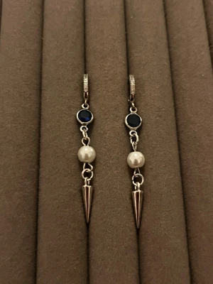Silvriga örhängen med pärlor och blå sten - Säljer ett par unika silvriga örhängen med blå sten upptill, vit pärla i mitten och en spetsig metallisk detalj längst ner. Örhängena har en modern och edgy design som sticker ut. Perfekta för dig som gillar att mixa klassiskt med trendigt.