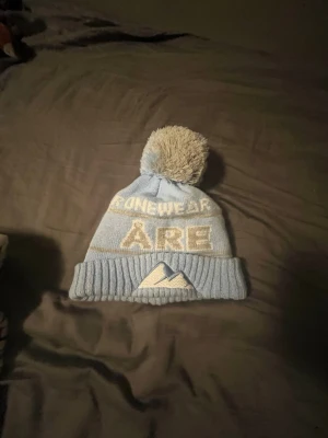 Ljusblå Åre mössa med tofs - Säljer en ljusblå stickad mössa från OneWear med stor tofs på toppen. Mössan har texten 'ÅRE' och ett bergmotiv framtill, samt invävd text runtom. Perfekt för kalla dagar och snygg till vinterlooken. 