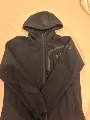 Svart hoodie från Under Armour - Snygg svart hoodie från Under Armour med dragkedja framtill och huva. Hoodien har diskreta loggor på bröstet och huvan. Perfekt för en avslappnad stil.