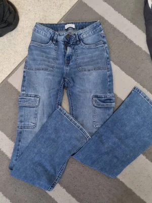 Blå cargobyxor från Lindex - Säljer ett par blå jeans från Lindex. Byxorna har utsvängda ben, stora fickor på sidorna och fram, samt klassiska bakfickor. 