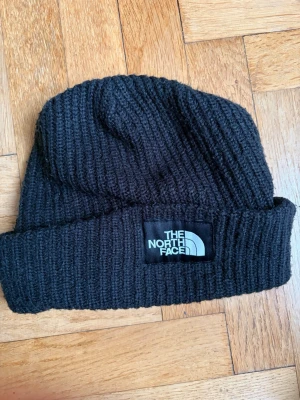 Svart ribbstickad barnmössa The North Face - Säljer en svart ribbstickad mössa från The North Face med uppvikt kant och tydlig logotyp framtill. Klassisk beanie-modell i mjukt och värmande material, perfekt för kyliga dagar. Enkel och stilren design som passar till det mesta.