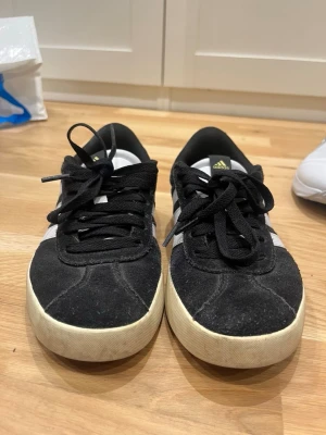 Svarta Adidas sneakers med vita detaljer - Säljer ett par svarta Adidas sneakers med vita detaljer och klassiska tre ränder på sidan. Skorna har svart mockaliknande material på ovandelen, vita sulor och svarta skosnören. Perfekta för en avslappnad streetstyle-look.