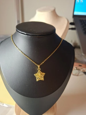 Guldigt halsband med stjärnhänge - Snyggt halsband i guldfärgad metall med ett stjärnformat hänge. Hänget har ett unikt, öppet mönster med detaljerade linjer som ger en cool och modern känsla. Perfekt accessoar för att lyfta din outfit med lite extra bling.