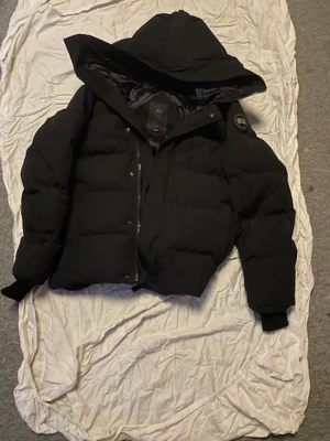 Svart Canada Goose Pufferjacka – Varm & Stilren Patch - Säljer en exklusiv svart Canada Goose pufferjacka med rymlig huva och quiltad design. Jackan har den ikoniska patchen på ärmen och ett elegant svart innerfoder. Perfekt isolering för kalla vinterdagar utan att kompromissa med stilen – en tidlös och eftertraktad streetwear-look som kombinerar funktion och fashion.