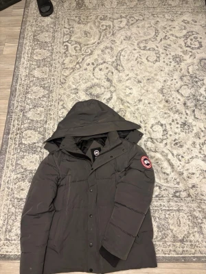 Mörkgrå dunjacka från Canada Goose - Säljer en mörkgrå dunjacka från Canada Goose med huva och klassisk logotyp på ärmen. Jackan har en stilren design med knappar och dragkedja framtill. Perfekt för kalla vinterdagar och riktigt varm tack vare dunfyllningen.
