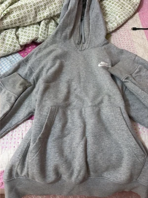 Grå hoodie från Nike med huva - Klassisk grå hoodie från Nike med vit logga på bröstet. Hoodien har stor känguruficka framtill, ribbade muddar och en rymlig huva med dragsko. Tillverkad i mjukt sweatshirtmaterial som är perfekt för chill dagar.