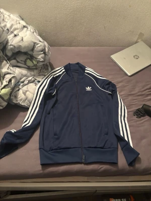 Blå Adidas track jacket - Snygg blå track jacket från Adidas med klassiska vita ränder längs ärmarna och logga på bröstet. Jackan har dragkedja framtill, ribbade muddar och hög krage. Perfekt för en sportig och avslappnad look.