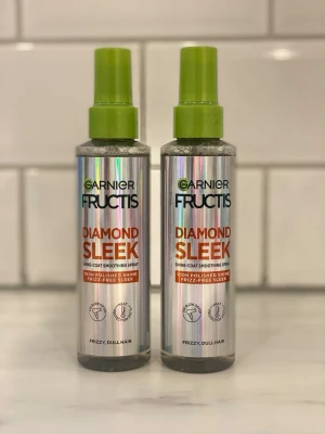 Garnier Fructis Diamond Sleek spray - Två flaskor Garnier Fructis Diamond Sleek Shine-Coat Smoothing Spray för frissigt och glanslöst hår. Flaskorna är silverfärgade med gröna lock och har text i svart och orange. Ger 100% polerad glans och slät finish.
