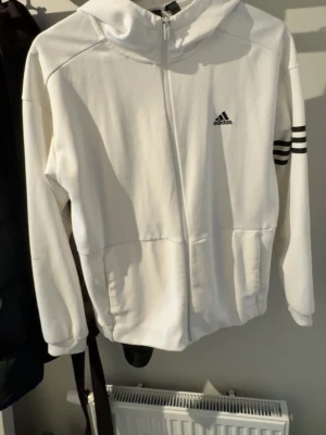 Vit hoodie från Adidas med dragkedja - Säljer en vit hoodie från Adidas med dragkedja framtill och svart Adidas-logga på bröstet. Tröjan har huva, ribbade muddar och tre svarta ränder på ena ärmen. Tillverkad i mjukt sweatshirtmaterial, perfekt för en sportig och avslappnad stil.