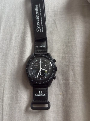 Omega x Swatch Speedmaster klocka svart - Snygg svart Omega x Swatch Speedmaster MoonSwatch med svart textilband och vita detaljer. Klockan har tachymeter på bezeln, flera urtavlor och tydliga loggor på bandet. Perfekt för dig som gillar klassisk men modern stil.