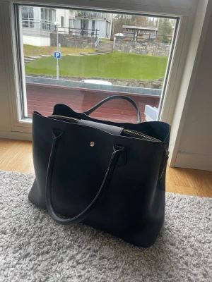 Svart stor handväska med gulddetaljer - (Handväskan ingår gratis i köpet!!)Snygg svart handväska i slätt konstläder med två långa handtag och guldfärgad dragkedja. Väskan har en stilren, rymlig design och en liten guldfärgad knappdetalj på framsidan. Perfekt för dig som vill ha plats för allt du behöver under dagen.