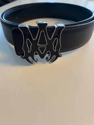 Svart Amiri bälte med metallspänne - Snyggt svart Amiri bälte med ett stort metallspänne i silverfärg. Bältet har en stilren och modern look, perfekt för att lyfta vilken outfit som helst. Tillverkat i skinn med detaljerad söm längs kanten och ett spänne som verkligen sticker ut.