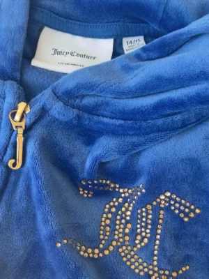 Blå Juicy Couture hoodie i velour - Säljer en blå hoodie från Juicy Couture i mjuk velour med dragkedja och huva. Guldiga detaljer som JC-logga i strass framtill och Juicy Couture-tryck på ryggen. Klassisk J-formad dragkedja. Perfekt för en trendig och avslappnad look.
