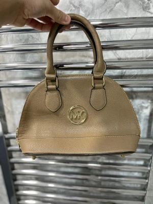 Beige handväska från Michael Kors - Snygg beige handväska från Michael Kors i strukturerat läder med guldfärgad MK-logga framtill. Väskan har två stadiga handtag och dragkedja upptill. Perfekt storlek för det viktigaste och en stilren look till din outfit.