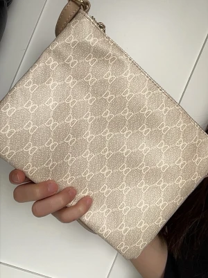 Beige väska - Säljer en beige axelväska från Don cos Donna med justerbar rem och guldfärgade detaljer. Väskan har ett mönster med Don Donnas logga över hela ytan och stängs med dragkedja. Tillverkad i tåligt konstläder som ger en stilren look.