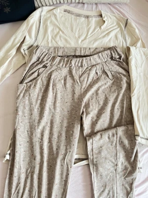 Beige blommig pyjamas med långärmad tröja - Mysig pyjamas med en cremevit långärmad tröja med v-ringning och kontrastsömm alpine detaljer. Byxorna är beige med små blommönster, har resår i midjan och sidofickor. Perfekt för chillkvällar hemma.