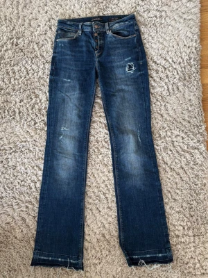Blå bootcut jeans från Guess - Säljer ett par mörkblå bootcut jeans från Guess med slitna detaljer. Jeansen har knäppning fram, med fina knapp detaljer. Materialet är stretchigt denim som sitter snyggt och bekvämt. De är långa och passar mig som är 178.                  Storlek: 25, Innerbenslängd: 88 cm