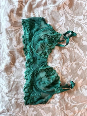 Två bhar i spets och blommigt mönster - Säljer två bhar: en smaragdgrön bh i transparent spets med broderade blommor och dekorativa band, samt en cremefärgad bh med småblommigt mönster, spetsdetaljer och rosetter. Båda har reglerbara axelband och bygel för extra stöd.