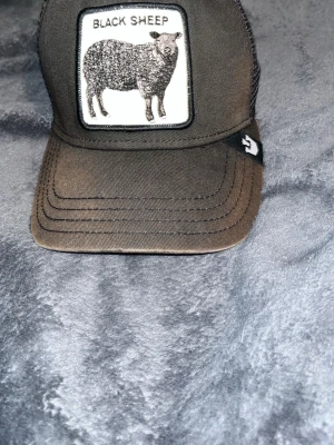 Svart truckerkeps Black Sheep - Säljer en svart truckerkeps från Goorin Bros med ett broderat patch framtill med texten 'Black Sheep' och en bild på ett svart får. Kepsen har mesh baktill för extra ventilation och justerbar snapback. Perfekt för dig som vill sticka ut. Lite som blekt brun aktig 