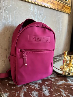 Vinröd ryggsäck från Gymshark - En mycket fin liten ryggväska från Gymshark. Ry cross har ett stort huvudfack, en framficka med dragkedja och justerbara axelband. Tillverkad i slitstarkt tyg med Gymshark-log mis lastat på framsidan. Perfekt för skola eller träning.