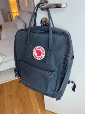 Blå Fjällräven Kånken ryggsäck - Klassisk svart Fjällräven Kånken ryggsäck i slitstarkt material med ikonisk röd logga framtill. Ryggsäcken har ett stort huvudfack, framficka med dragkedja och två sidofickor. Praktiska handtag upptill och justerbara axelremmar.