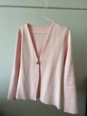 Ljusrosa kofta med guldknapp - Söt ljusrosa kofta med en dekorativ guldknapp framtill. Ko soppen har v-ringning, långa ärmar och är tillverkad soppen i ett mj restickat material. Perfekt för dig som gillar en enkel men elegant stil.