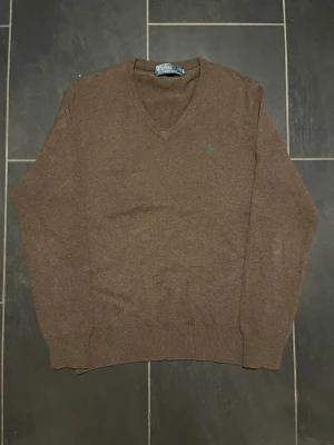 Brun V-ringad tröja Polo Ralph Lauren - Klassisk brun V-ringad tröja från Polo Ralph Lauren med broderad grön logga på bröstet. Tröjan är stickad i mjukt material och har ribbade muddar vid ärmslut och nederkant. Perfekt för lager-på-lager-stil.