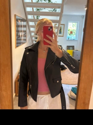 Svart skinnjacka - Cool svart skinnjacka med klassisk bikerstil, silverfärgade dragkedjor och knappar. Jackan har ett bälte i midjan, snedställda fickor och breda slag. Perfekt för dig som vill ha en edgy look och gillar detaljer.       Lite skav på insidan kragen men det syns inte när man har på sig den.