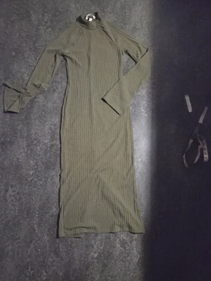 SOM NY Olivgrön ribbad långklänning stl XS  - Snygg olivgrön långklänning med ribbad struktur och lång ärm. STL XD Klänningen har lite högre hals och en slits nedtill på ena sidan för extra stil. Perfekt för dig som gillar en enkel men trendig look. LÄNGD FRÅN AXEL OCH NER 114 CM 