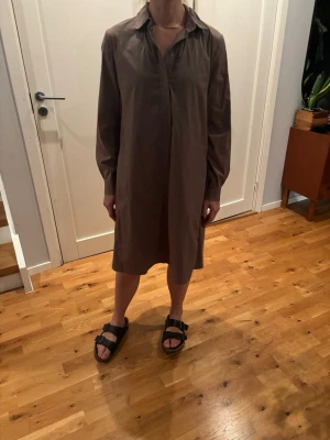 Brun skjortklänning med lång ärm - En enkel och stilren brun skjortklänning med krage och långa ärmar. Klänningen har A-linjeformad passform och slutar strax ovanför knäna. Tillvererkad ett mjukt bomullsliknande material, perfekt för en avslappnad men snygg look.