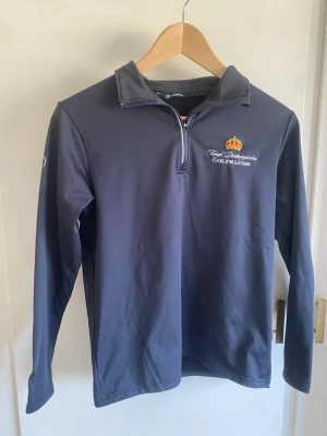 Mörkblå golftröja från Abacus - Mörkblå långärmad golftröja från Abacus med halv dragkedja och broderad logga för finger Boströmska Golfklubb på bröstet. Tillverkad i ett stretchigt funktionsmaterial som passar perfekt för sportiga aktiviteter. trailing logga på ryggen.