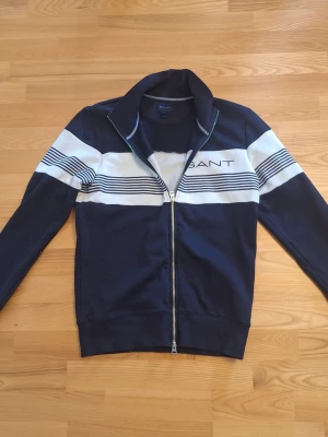Marinblå ziptröja från Gant - Snygg marinblå zip, sweatshirttröja från Gant med vita och blå ränder över bröstet och armarna. Tröjan har hög krage, dragkedja framtill och Gant-logga broderad på bröstet. ]Perfekt för en sportig och avslappnad stil.