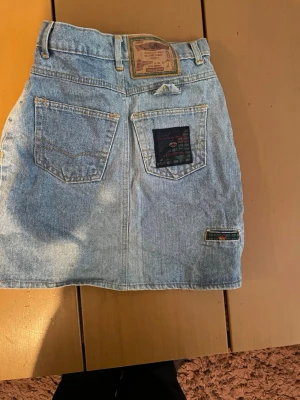 Ljusblå jeanskjol från Levi's - Säljer en klassisk ljusblå jeanskjol från Levi's med fem fickor, bälteshällor och knappstängning fram. Kjolen har en rak passform och detaljerade Levi's-lappar på bakfickan och baktill i midjan. Perfekt till sneakers eller boots!