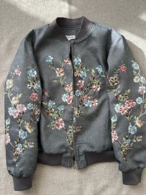 Grå bomberjacka med blommigt mönster - Supersnygg grå bomberjacka från Siesta med broderade blommor med pärlor i pastellfärger över hela jackan. Jackan har ribbade muddar, dragkedja framtill och en riktigt unik look med sina färgglada detaljer. Perfekt för dig som vill sticka ut med en cool och trendig stil.passar S M L